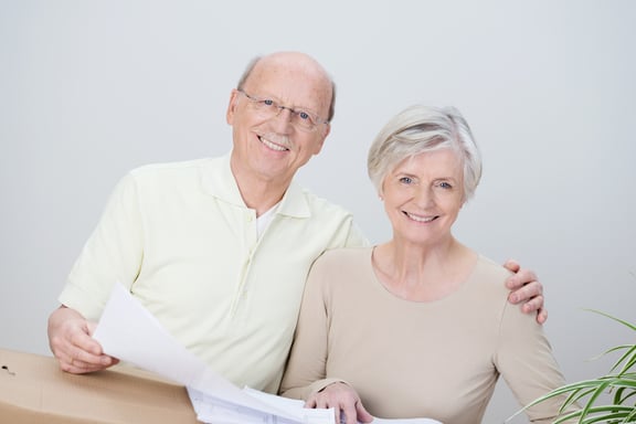 Smiling affectionate senior couple standing with the husbands arm around his wife looking at documents resting on a brown cardboard carton as they plan their move to a new home-4